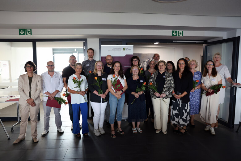 Gruppenfoto von 18 Personen bei einer Ehrung, mehrere halten Rosen. Sie stehen vor einer Stellwand mit Sachsen-Logo.