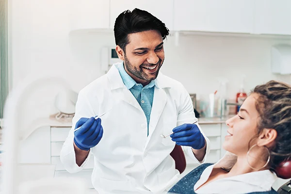 Arzt blick lächelnd auf Patientin im Stuhl, in der Hand hält er ein Dentalinstrument.