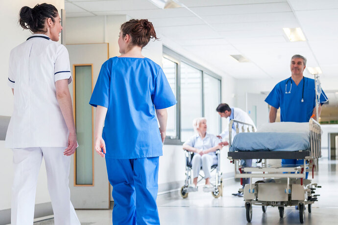 Medizinisches Personal auf Krankenhausflur: Pflegekräfte begleiten ein leeres Krankenhausbett, im Hintergrund sitzt eine Patientin im Rollstuhl.