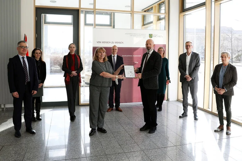 Neun Personen in Eingangshalle bei offizieller Übergabe, zwei Frauen in der Mitte halten Dokument, im Hintergrund Roll-up-Banner mit Sachsen-Wappen.