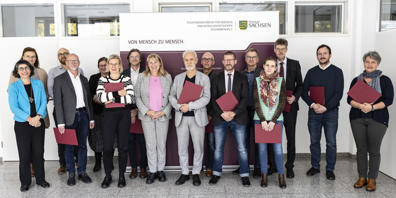 Gruppe von Menschen mit roten Mappen steht vor Banner des Sächsischen Staatsministeriums für Soziales in hellem Raum.
