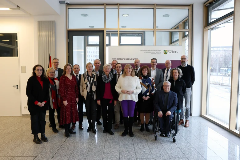 Gruppe von Menschen steht vor Banner des Sächsischen Staatsministeriums in hellem Raum mit Fenstern, Person im Rollstuhl vorn.