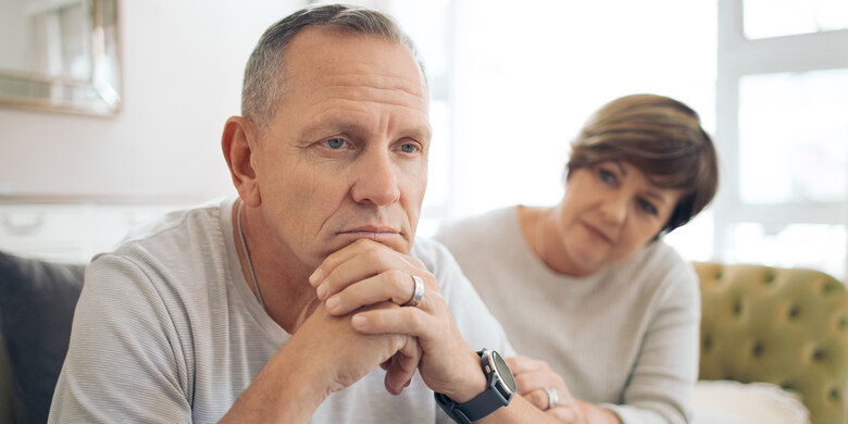 Mann sitzt nachdenklich im Vordergrund, Frau im unscharfen Hintergrund. Häusliche Gesprächssituation.