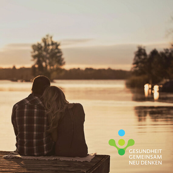 Ein Paar sitzt innig am Ufer eines Sees und blickt gemeinsam auf das spiegelnde Wasser im ruhigen Abendlicht.