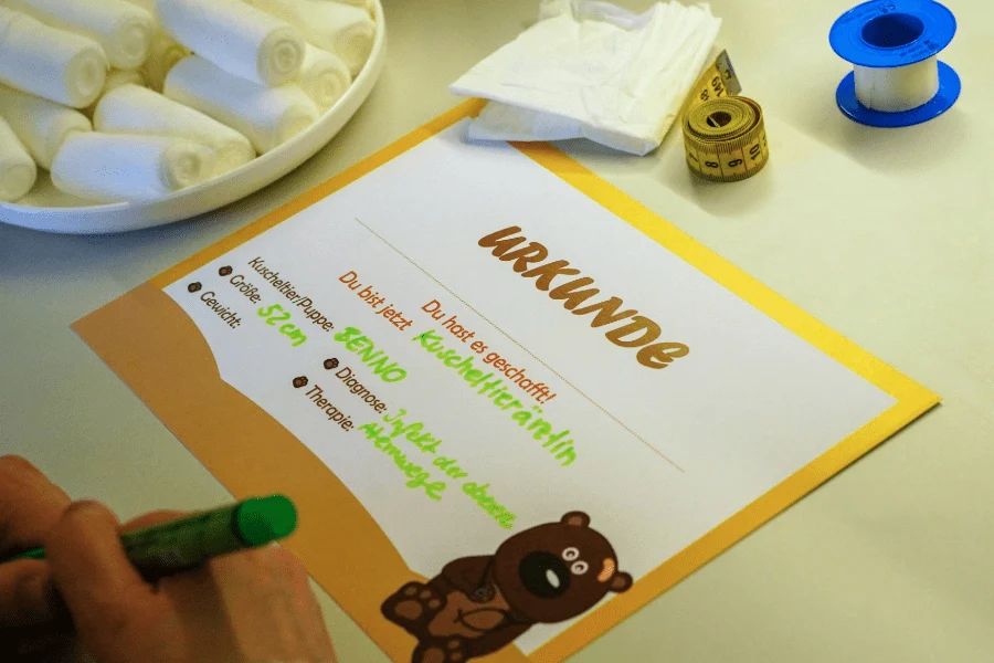 Informationsmaterial zur Wundversorgung: Kind malt auf ein Plakat mit bunten Wundarten wie Biene, Schnitt oder Schürfwunde. Verbandmaterial liegt bereit.