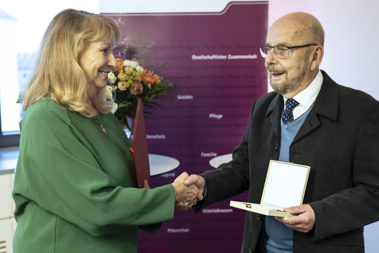 Frau in grünem Blazer und Mann im Anzug geben sich die Hand bei offizieller Urkundenübergabe vor Stellwand.