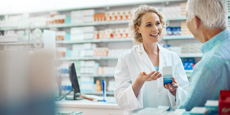 Apothekerin in weißem Kittel berät Kundin in einer Apotheke und überreicht Medikamentenpackung.