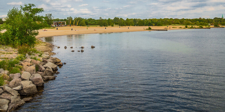 Blick auf Badesee mit Sandstrand, steinigem Ufer, grünen Bäumen und Badegästen unter bewölktem Himmel.