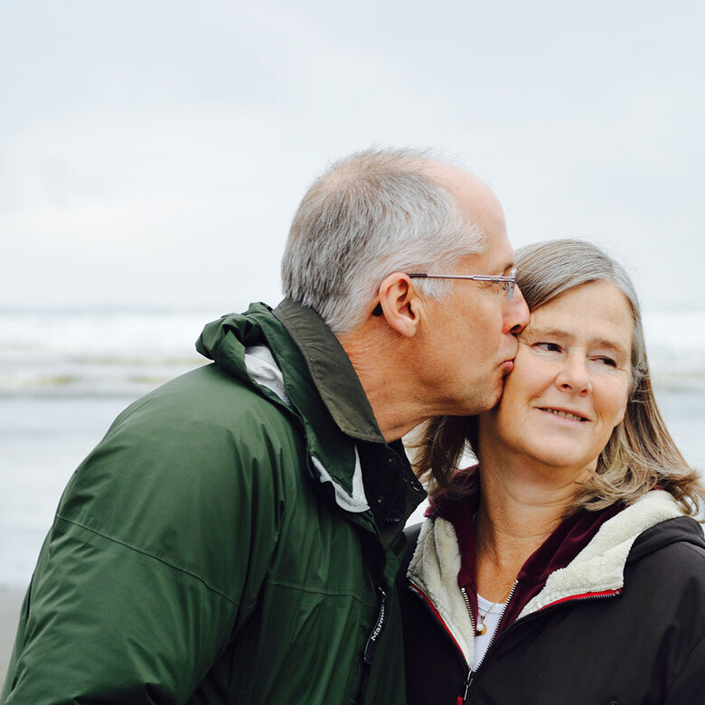 Mann mit grauer Brille und grüner Jacke küsst Frau auf die Wange am Strand, beide mittleren Alters, sie lächelt.