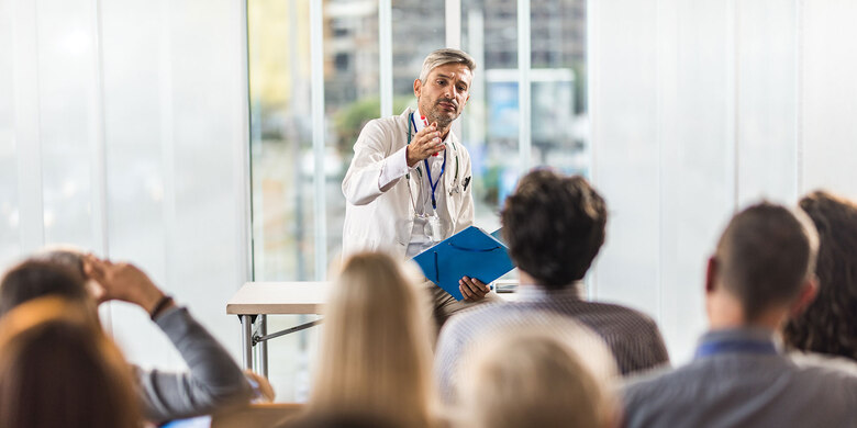 Arzt im Kittel mit Stethoskop hält blaue Mappe und spricht vor sitzendem Publikum in hellem Raum.