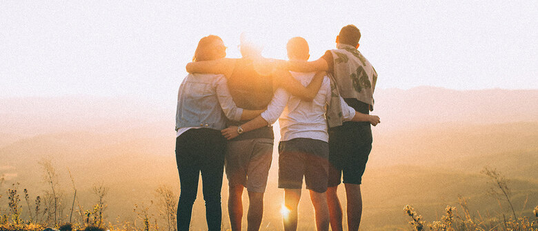 Vier Personen stehen eng umarmt auf einem Hügel mit Blick in die Abendsonne. Die Rücken sind zugewandt, das Sonnenlicht erzeugt eine warme, freundschaftliche Atmosphäre im Freien.