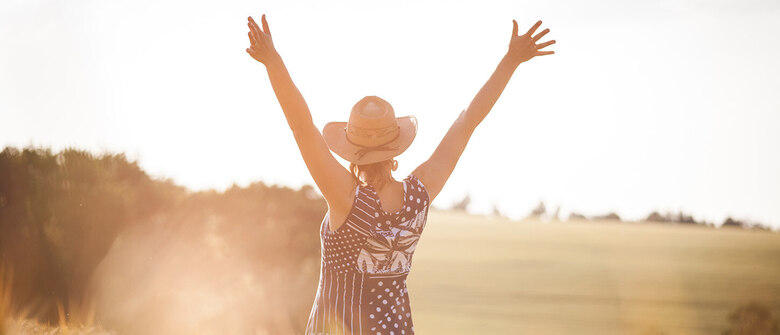 Eine Frau mit Strohhut steht im Sonnenlicht auf einem Feld, die Arme weit nach oben ausgestreckt. Sie trägt ein ärmelloses Kleid mit hellem Muster und wirkt fröhlich und frei in einer sommerlichen Landschaft.