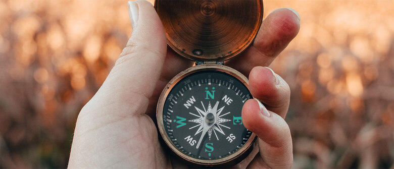 Eine Hand hält einen geöffneten Kompass vor unscharfem, beige-braunem Naturhintergrund. Die Kompassnadel weist nach Norden. Das Motiv vermittelt Orientierung und Richtung in einer natürlichen Umgebung.