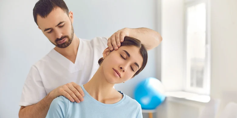 Therapeut in weißem Hemd dehnt Nacken einer Frau in hellblauem Shirt in hellem Raum, Behandlungsszene.