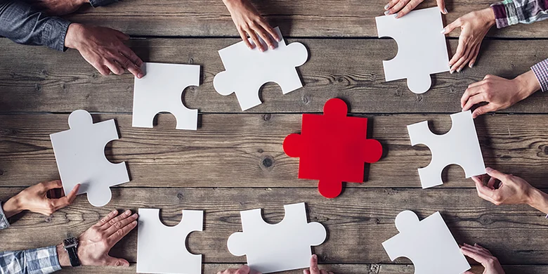 Hände halten weiße Puzzleteile um rotes Puzzleteil auf Holztisch, Symbol für Teamarbeit und Zusammenarbeit.