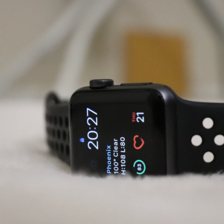 Schwarze Smartwatch mit Display zeigt Uhrzeit 20:27, Gesundheitsdaten und Symbole, schwarzes Sportarmband mit Löchern, unscharfer Hintergrund.