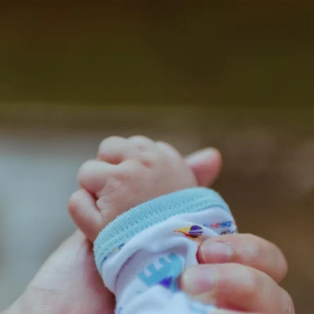 Eine erwachsene Hand hält sanft die kleine Hand eines Babys, das einen weißen Strampler mit buntem Muster trägt. Der Fokus liegt auf den Händen, der Hintergrund ist unscharf, die Szene wirkt beschützend und zärtlich.