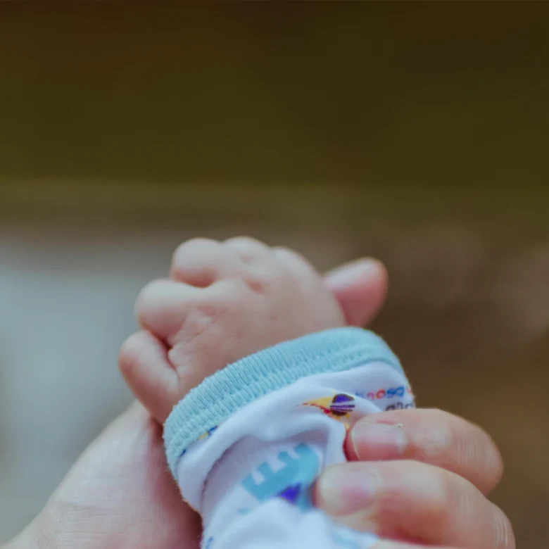 Eine erwachsene Hand hält sanft die kleine Hand eines Babys, das einen weißen Strampler mit buntem Muster trägt. Der Fokus liegt auf den Händen, der Hintergrund ist unscharf, die Szene wirkt beschützend und zärtlich.