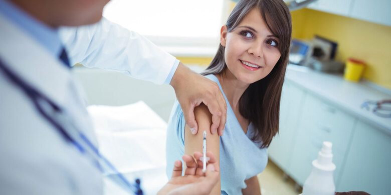 Ärztin impft lächelnde Frau in hellblauem Oberteil am Arm in heller Praxis.