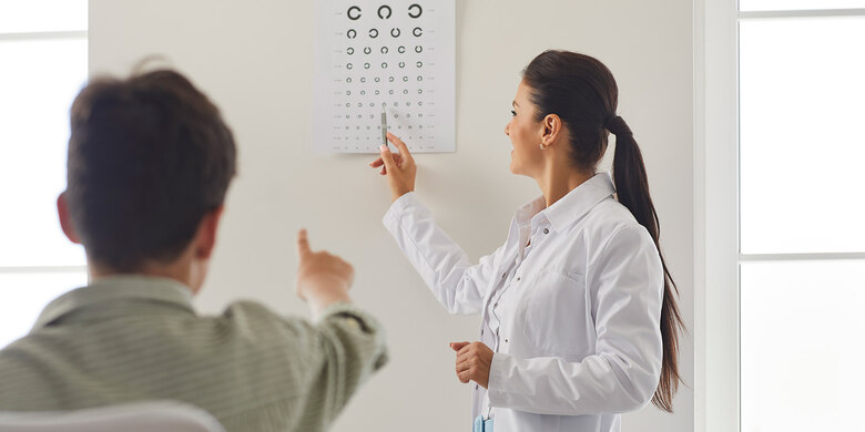 Ärztin in weißem Kittel zeigt mit Stab auf Sehtesttafel, Person sitzt davor in hellem Raum.