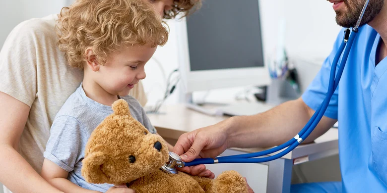 Arzt in blauer Kleidung untersucht Teddy mit Stethoskop, Kind mit lockigem Haar und Mutter schauen zu in heller Praxis.
