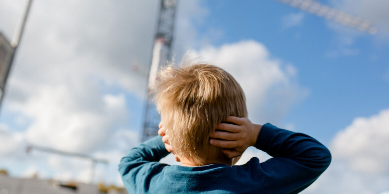 Kind mit blondem Haar hält Ohren zu vor unscharfem Hintergrund mit Windrad und blauem Himmel.
