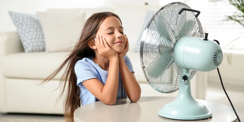 Mädchen in hellblauem Shirt sitzt vor türkisem Ventilator, hält beide Hände ans Gesicht, wehende Haare, heller Raum.