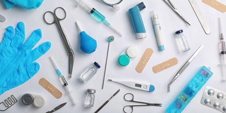 Medizinische Utensilien auf weißem Untergrund: blaue Handschuhe, Spritzen, Inhalatoren, Pflaster, Scheren, Thermometer, Pillendose und Tabletten.