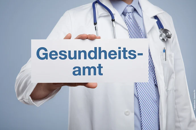 Ärztin im weißen Kittel mit Stethoskop hält Schild mit Text „Gesundheitsamt".