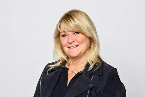 Porträt einer blonden Frau in dunkler Jacke vor hellem Hintergrund.