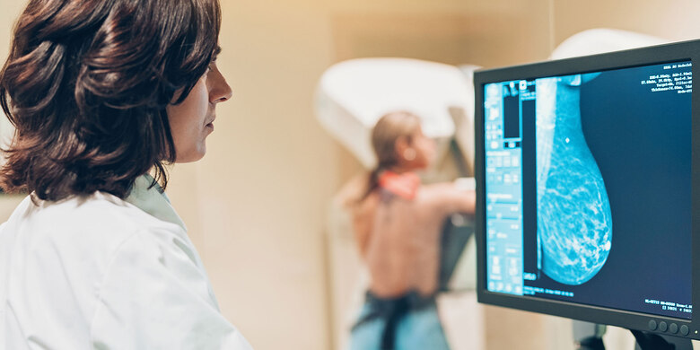 Ärztin betrachtet Mammografie-Aufnahme am Monitor. Patientin im unscharfen Hintergrund.