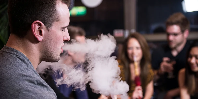 Mann raucht Shisha und stößt Dampfwolke aus. Soziale Umgebung mit Personen im unscharfen Hintergrund.