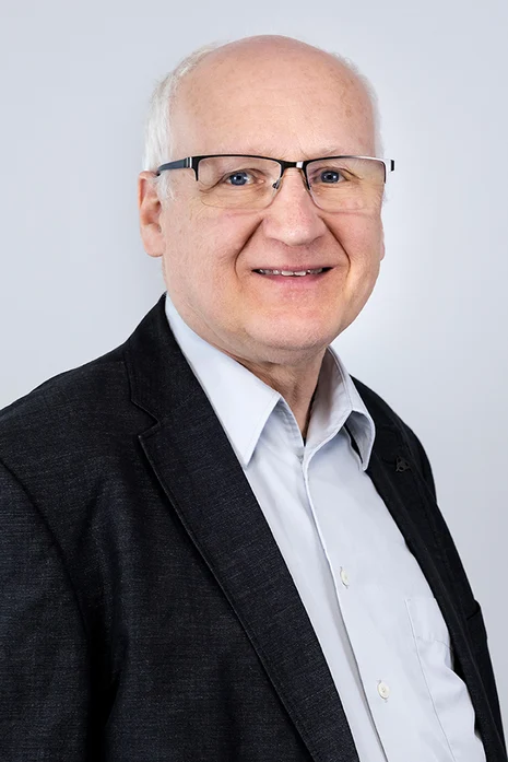 Dipl.-Med Stefan Mertens, Porträt eines älteren Mannes mit Brille in dunklem Sakko und weißem Hemd vor hellem Hintergrund.