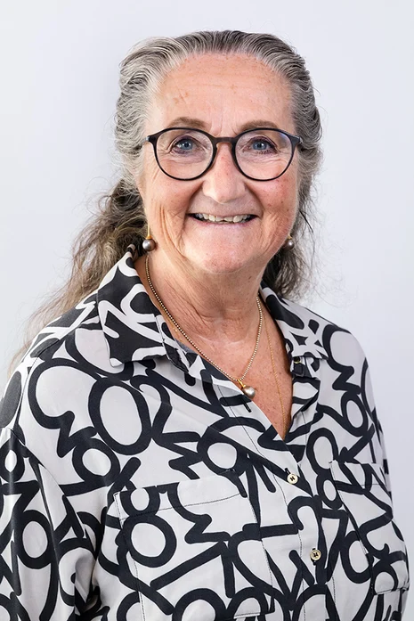 Dr. med. Patricia Klein, Porträt einer lächelnden älteren Frau mit Brille in schwarz-weiß gemusterter Bluse mit goldener Kette vor hellem Hintergrund.
