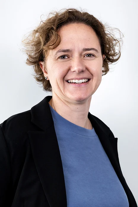 Dr. med. Friederike Neumann, Porträt einer lächelnden Frau mit lockigem braunem Haar in schwarzem Blazer über blauem Shirt vor hellem Hintergrund.