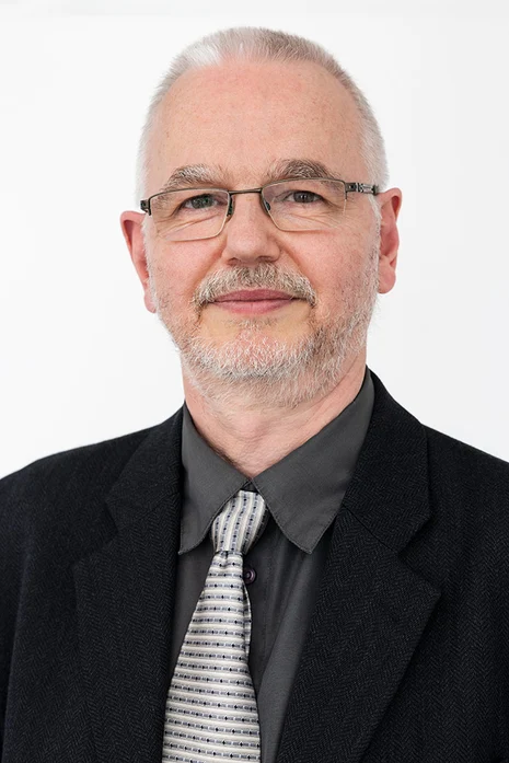 Dr. med. Thomas Grünewald, Porträt eines Mannes mit Brille und Bart in schwarzem Sakko, grauem Hemd und gestreifter Krawatte vor hellem Hintergrund.