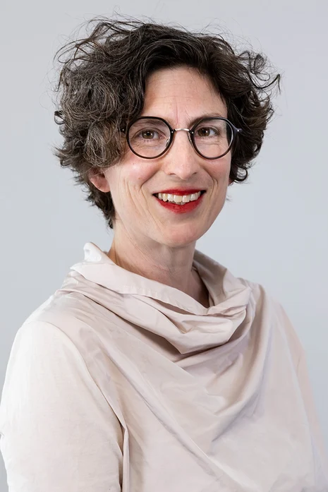 Prof. Dr. med. Birte Pantenburg, Porträt einer lächelnden Frau mit Brille und lockigem Haar in beiger Bluse mit Schal vor hellem Hintergrund.