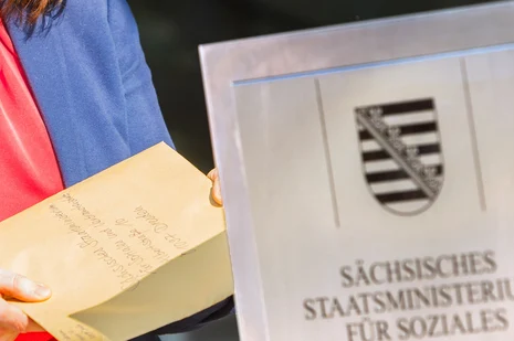 Person hält gelben Ordner vor Schild des Sächsischen Staatsministeriums für Soziales.