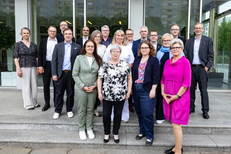 Gruppe von Personen vor Gebäude. Frauen und Männer in Anzügen und Kleidern stehen auf Stufen vor Glasfassade.