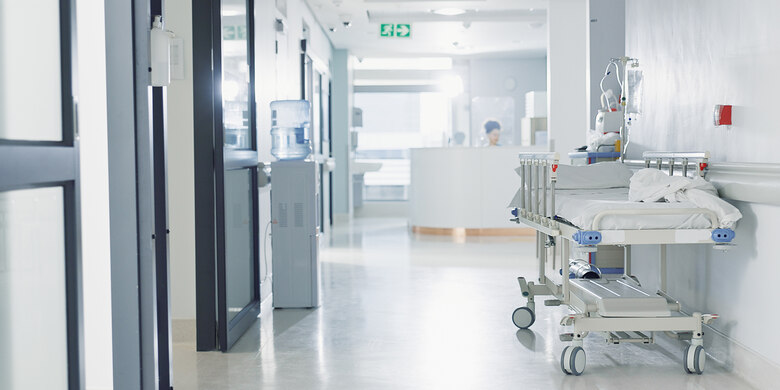 Krankenhausflur mit Patientenbett. Helles Interieur, Infusionsständer, Notausgangsschild, Personal im Hintergrund, medizinische Ausstattung.