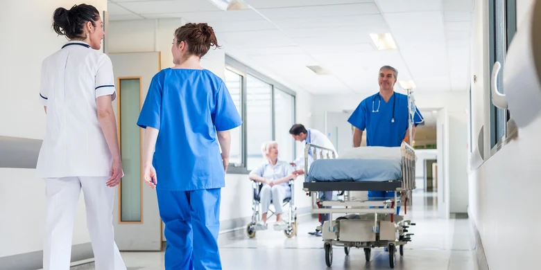 Medizinisches Personal auf Krankenhausflur: Pflegekräfte begleiten ein leeres Krankenhausbett, im Hintergrund sitzt eine Patientin im Rollstuhl.