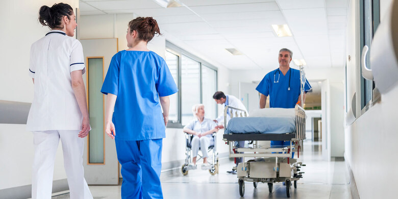 Medizinisches Personal auf Krankenhausflur: Pflegekräfte begleiten ein leeres Krankenhausbett, im Hintergrund sitzt eine Patientin im Rollstuhl.