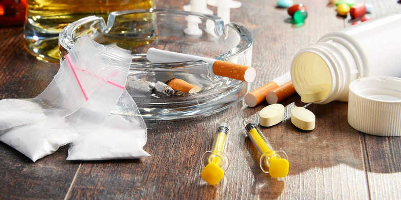 Darstellung von Suchtmitteln: Aschenbecher mit Zigaretten, Alkohol, Tabletten, Drogenparaphernalia mit Spritzen und weißem Pulver auf Holztisch.