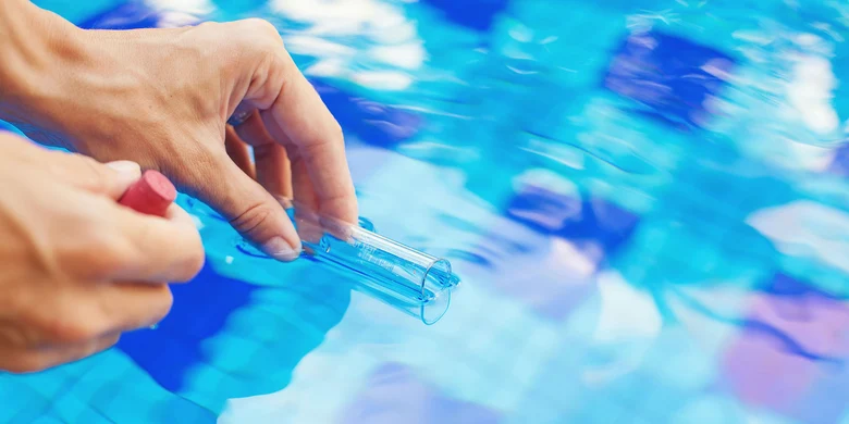 Hand entnimmt Wasserprobe über Schwimmbecken, symbolisiert umweltbezogenen Gesundheitsschutz und Überwachung der Badewasserqualität durch Gesundheitsämter.