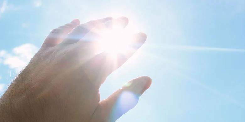 Hand greift nach Sonnenstrahlen vor blauem Himmel, symbolisiert UV-Strahlung und Notwendigkeit von Sonnenschutz zur Hautkrebsprävention.