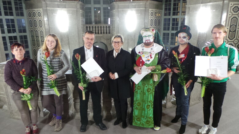 Verleihung der Sächsischen Ehrenmedaille für herausragende Leistungen im Kampf gegen HIV und AIDS 2016: Sieben Preisträger mit Urkunden und roten Rosen in Gebäude.