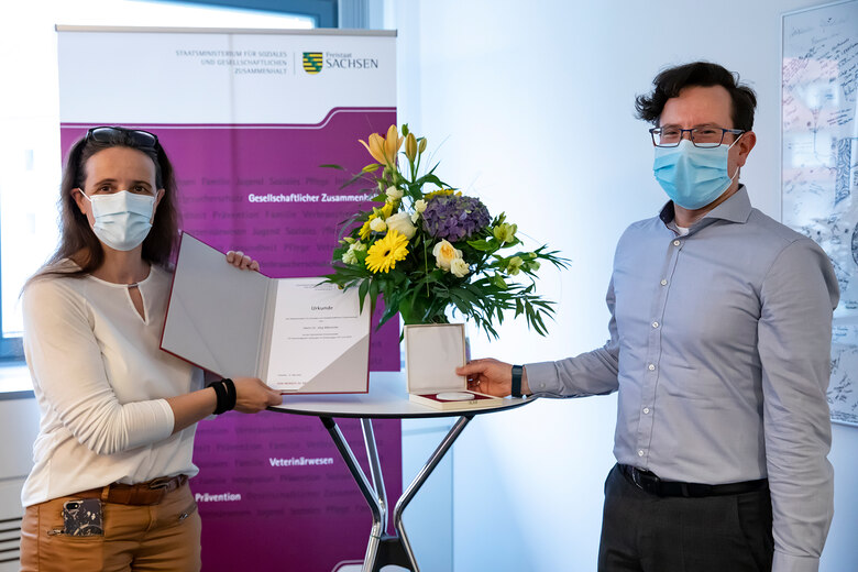 Verleihung der Sächsischen Ehrenmedaille für herausragende Leistungen im Kampf gegen HIV und AIDS 2019/2020: Zwei Personen mit Mund-Nasen-Schutz, Blumenstrauß und Medaille.