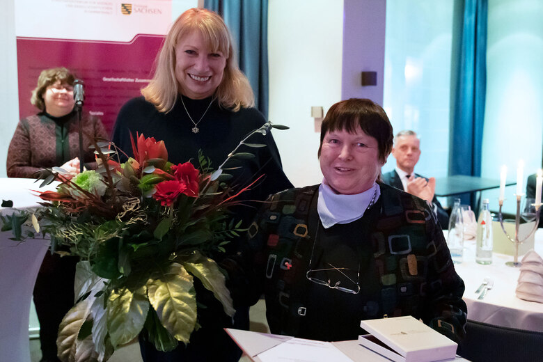 Verleihung der Sächsischen Ehrenmedaille für herausragende Leistungen im Kampf gegen HIV und AIDS 2022: Zwei Frauen mit Blumenstrauß und Urkunde am Tisch.